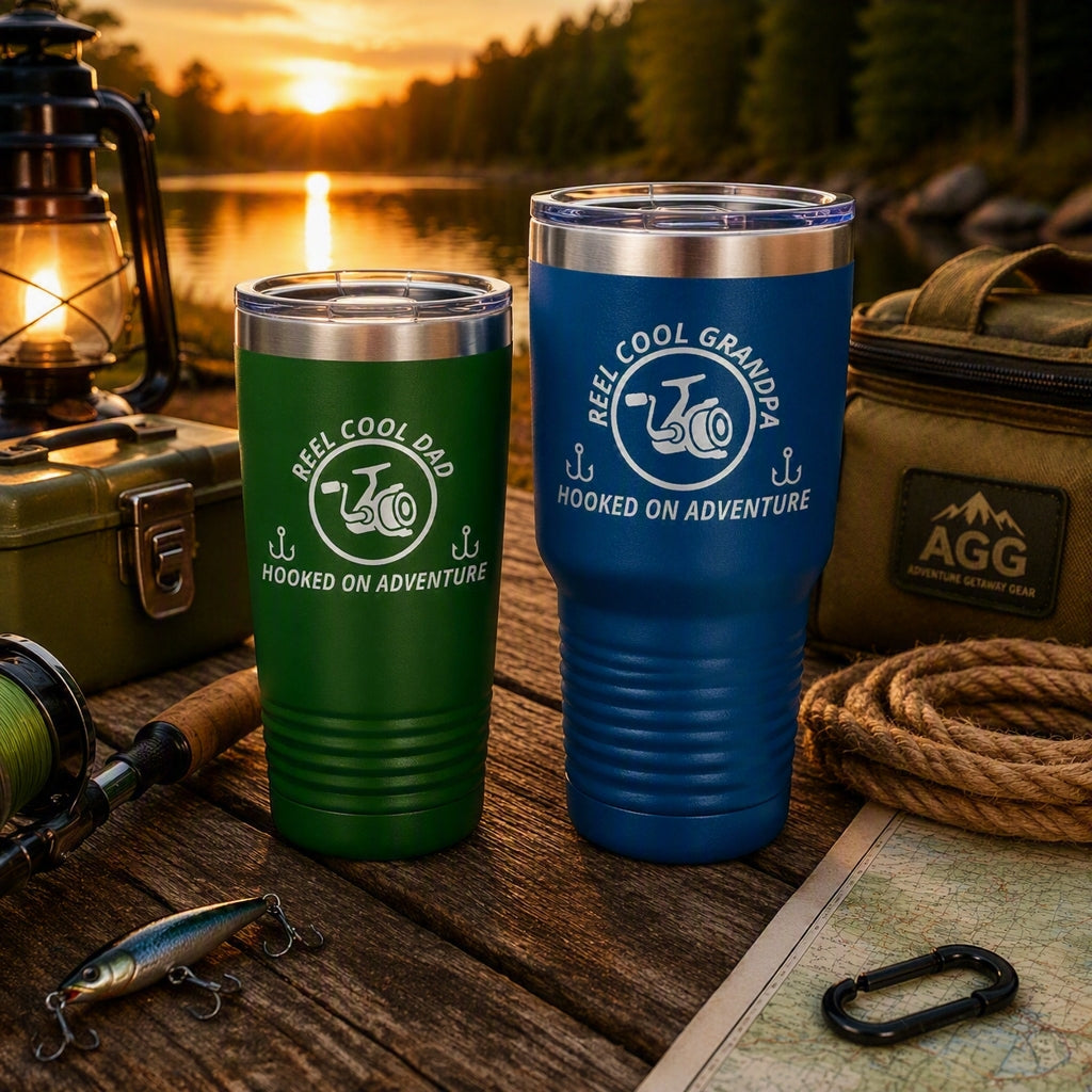 Green 20oz REEL COOL DAD tumbler and blue 30oz REEL COOL GRANDPA tumbler on a rustic lakeside table with fishing gear at sunset.