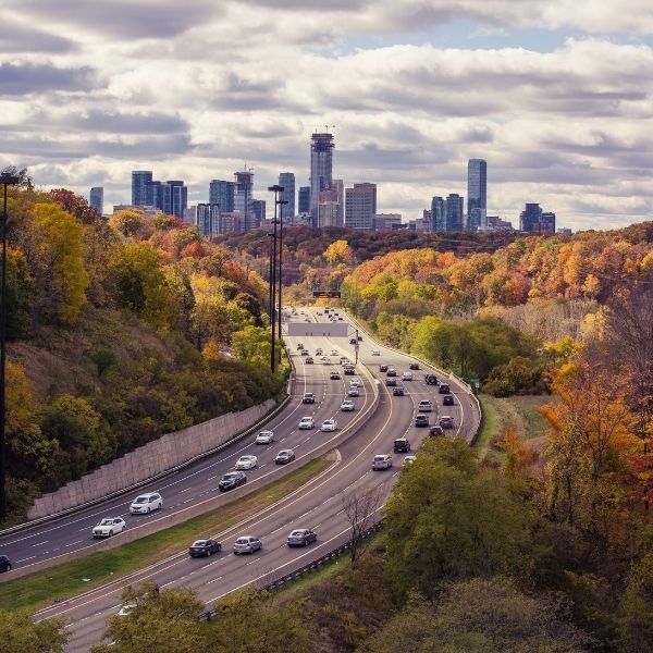 Cars on a curving highway through autumn trees with a city skyline in the distance.