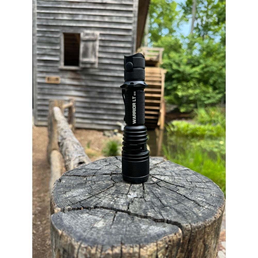 Powertac Warrior LT G5 flashlight standing upright on a weathered tree stump in front of a rustic wooden mill and creek.