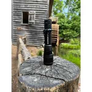 Powertac Warrior LT G5 flashlight standing upright on a weathered tree stump in front of a rustic wooden mill and creek.