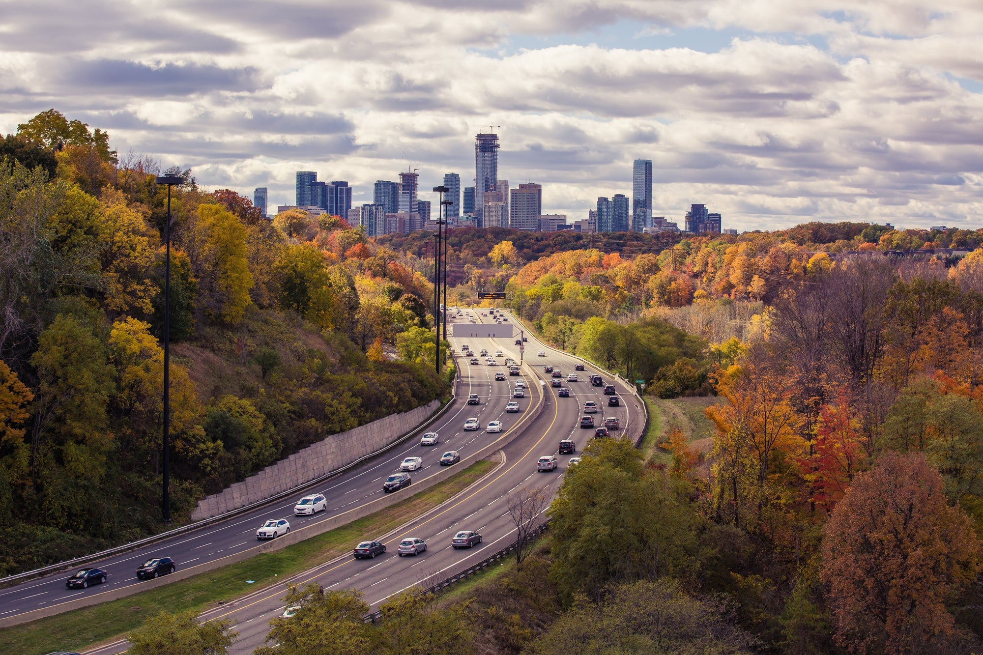 Cars on a curving highway through autumn trees with a city skyline in the distance.