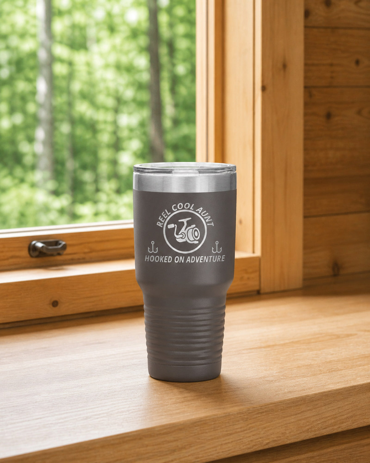 Pewter 30-ounce REEL COOL AUNT engraved tumbler on a bright wood cabin counter beside a window overlooking green forest.