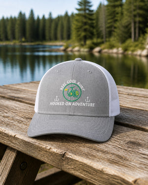Heather Grey and White REEL COOL AUNT embroidered trucker cap on a weathered wooden picnic table beside a pine-lined forest lake.