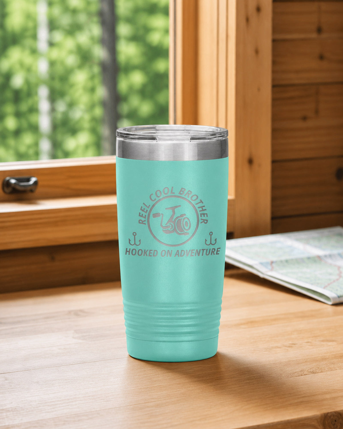 Teal 20-ounce REEL COOL BROTHER engraved tumbler on a bright wood cabin counter beside a window overlooking green forest.