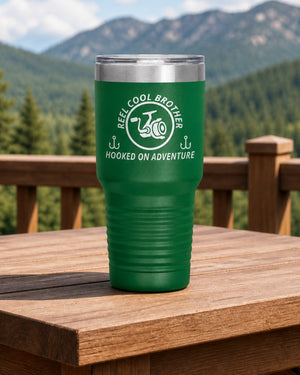 Green 30-ounce REEL COOL BROTHER engraved tumbler on a sturdy cabin porch table overlooking forested mountains.