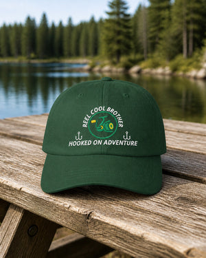Spruce REEL COOL BROTHER embroidered dad cap on a weathered wooden picnic table beside a pine-lined forest lake.