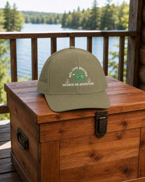 Loden REEL COOL BROTHER embroidered trucker cap on a cedar gear chest overlooking a pine-lined forest lake.