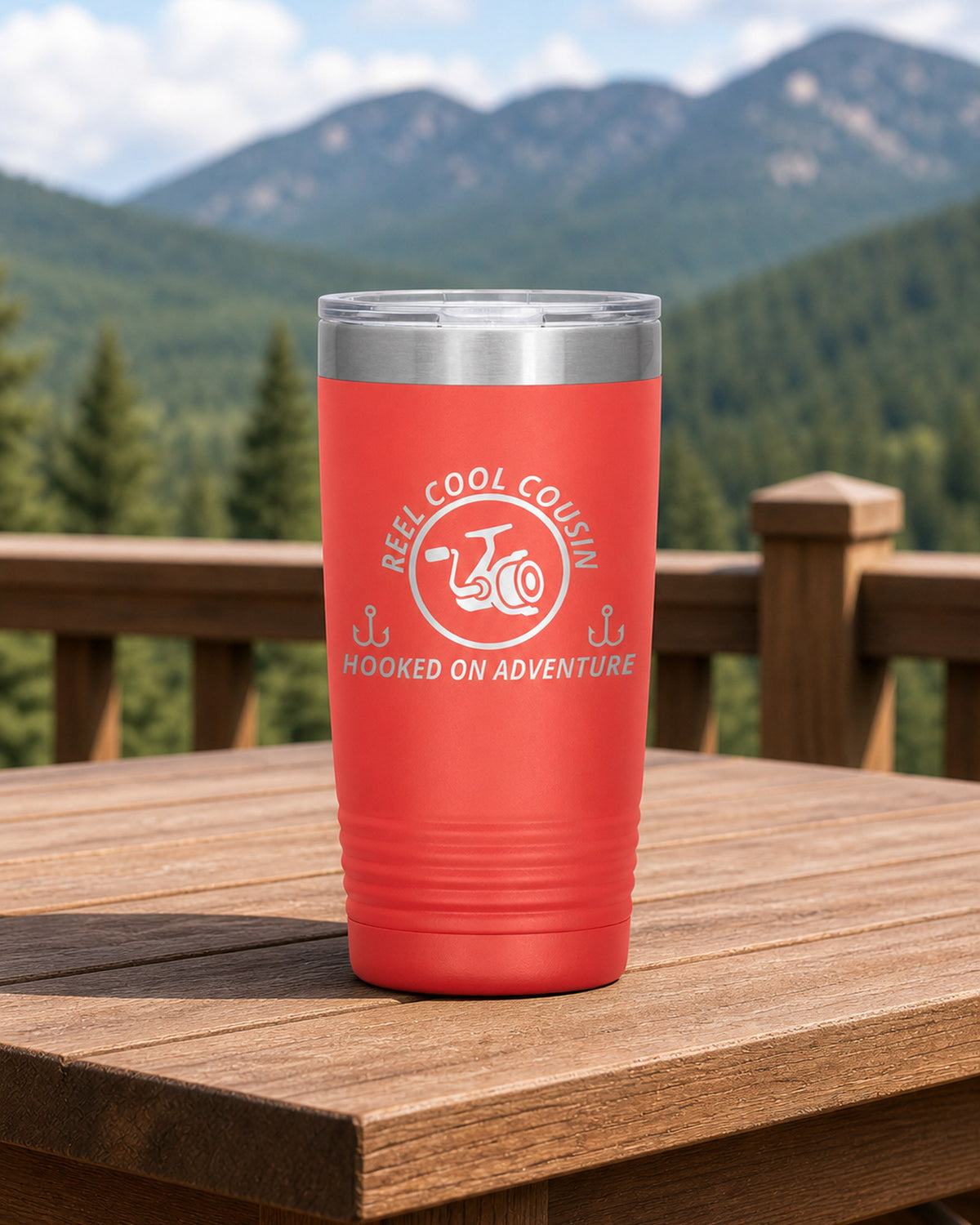 Coral 20-ounce REEL COOL COUSIN engraved tumbler on a sturdy cabin porch table overlooking forested mountains.