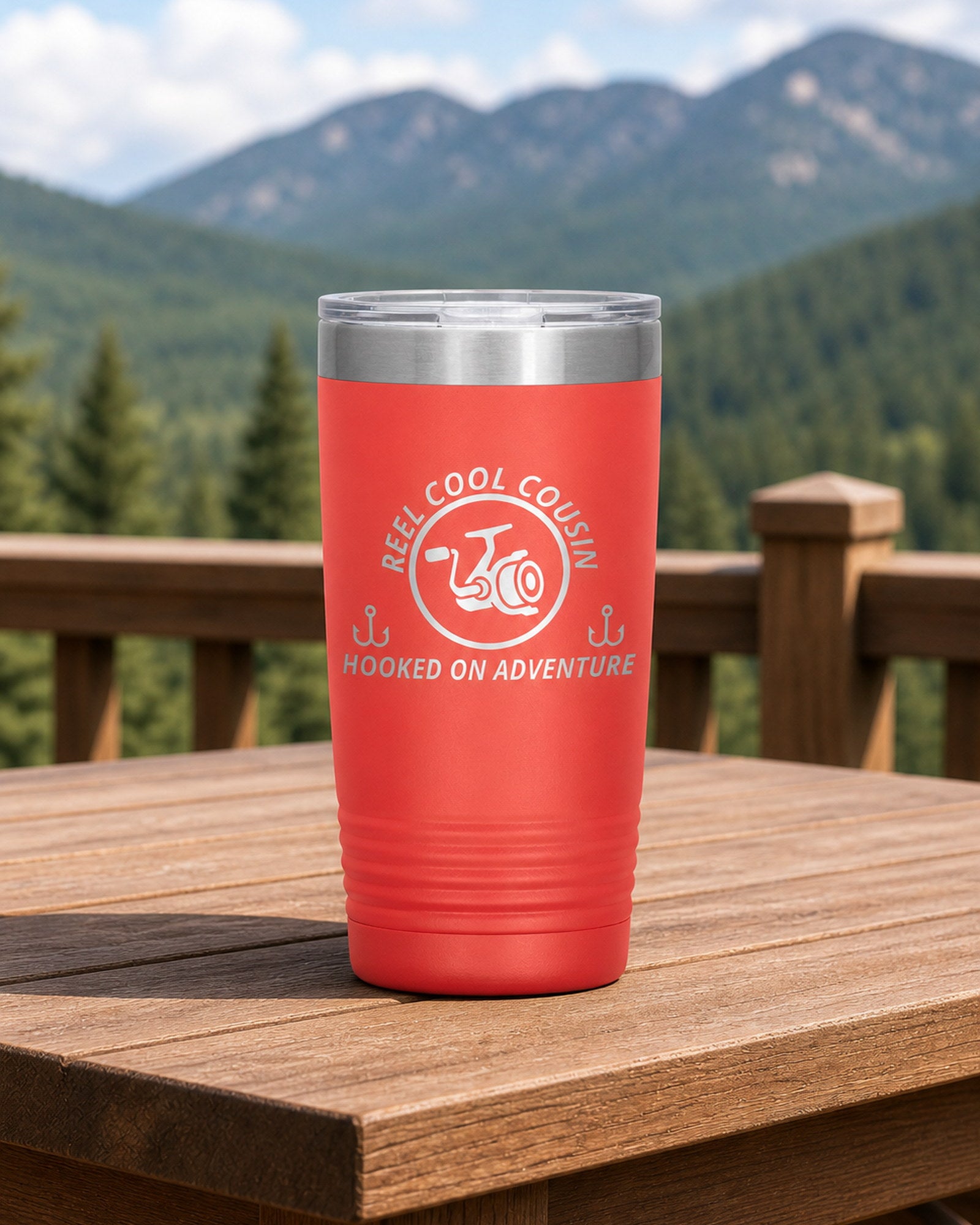 Coral 20-ounce REEL COOL COUSIN engraved tumbler on a sturdy cabin porch table overlooking forested mountains.