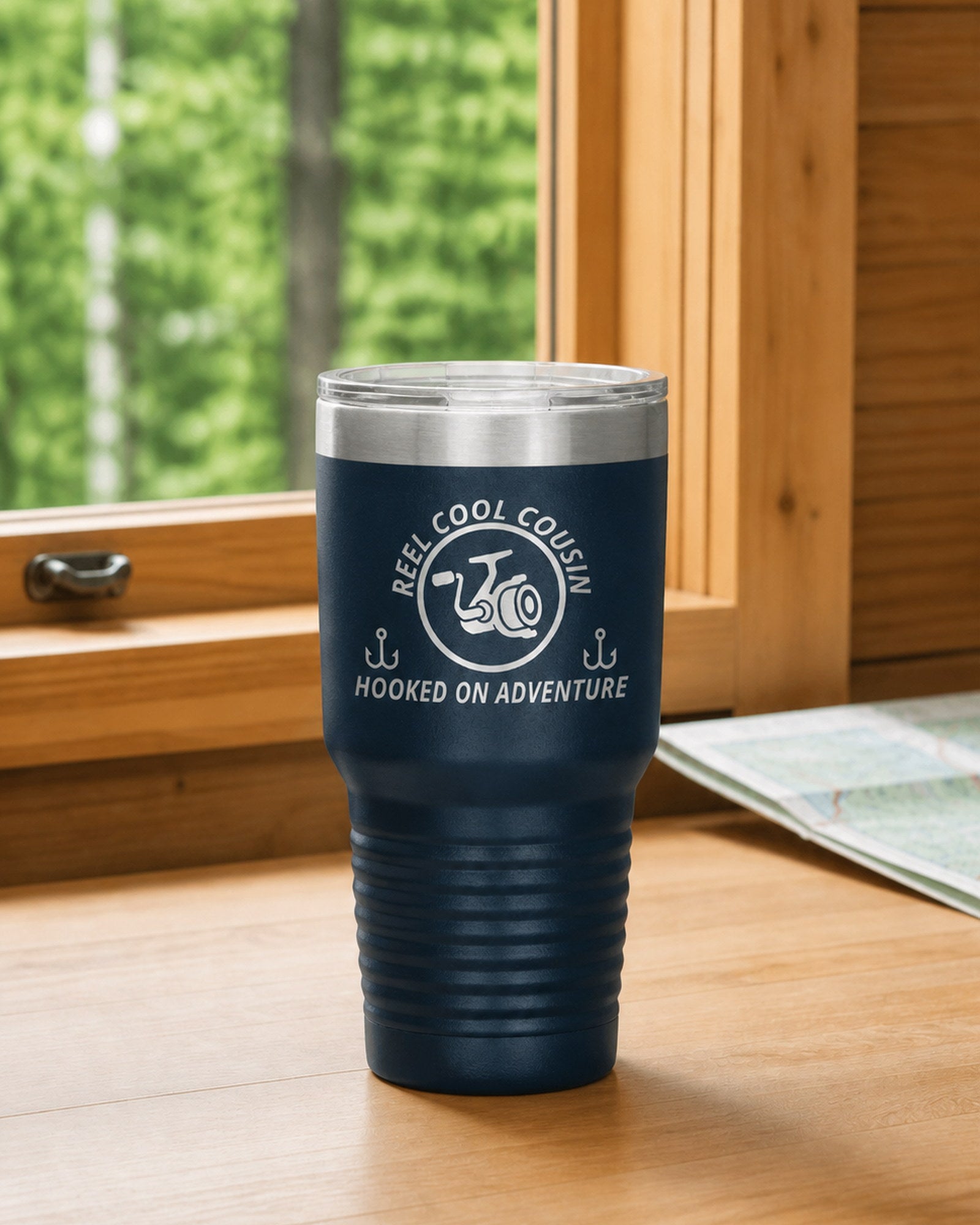 Navy 30-ounce REEL COOL COUSIN engraved tumbler on a bright wood cabin counter beside a window overlooking green forest.