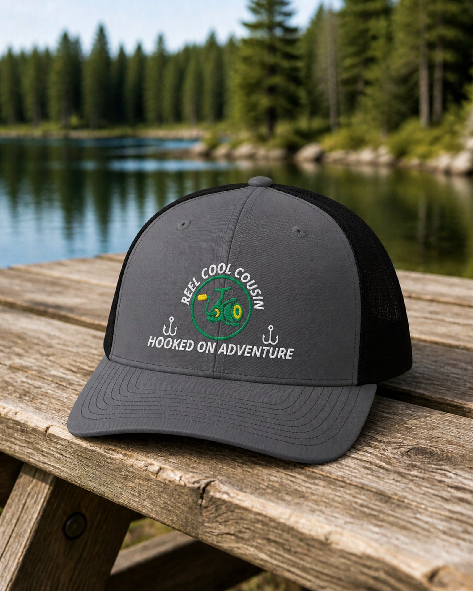 Charcoal and Black REEL COOL COUSIN embroidered trucker cap on a weathered wooden picnic table beside a pine-lined forest lake.