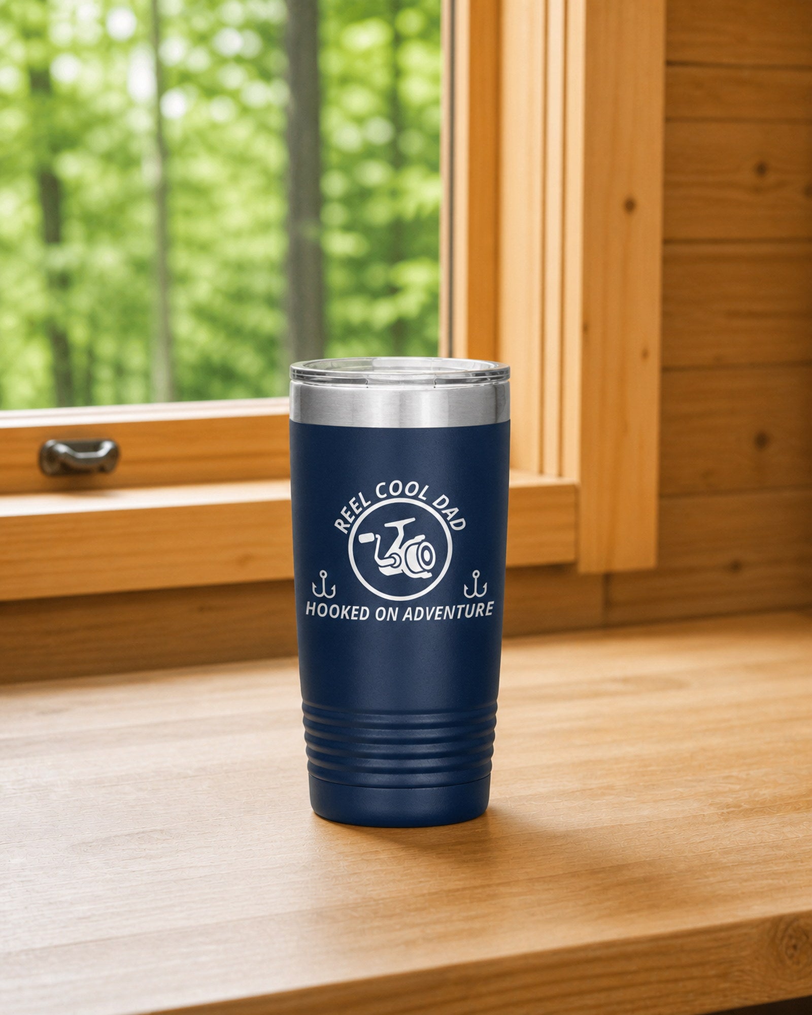 Navy 20-ounce REEL COOL DAD engraved tumbler on a bright wood cabin counter beside a window overlooking green forest.