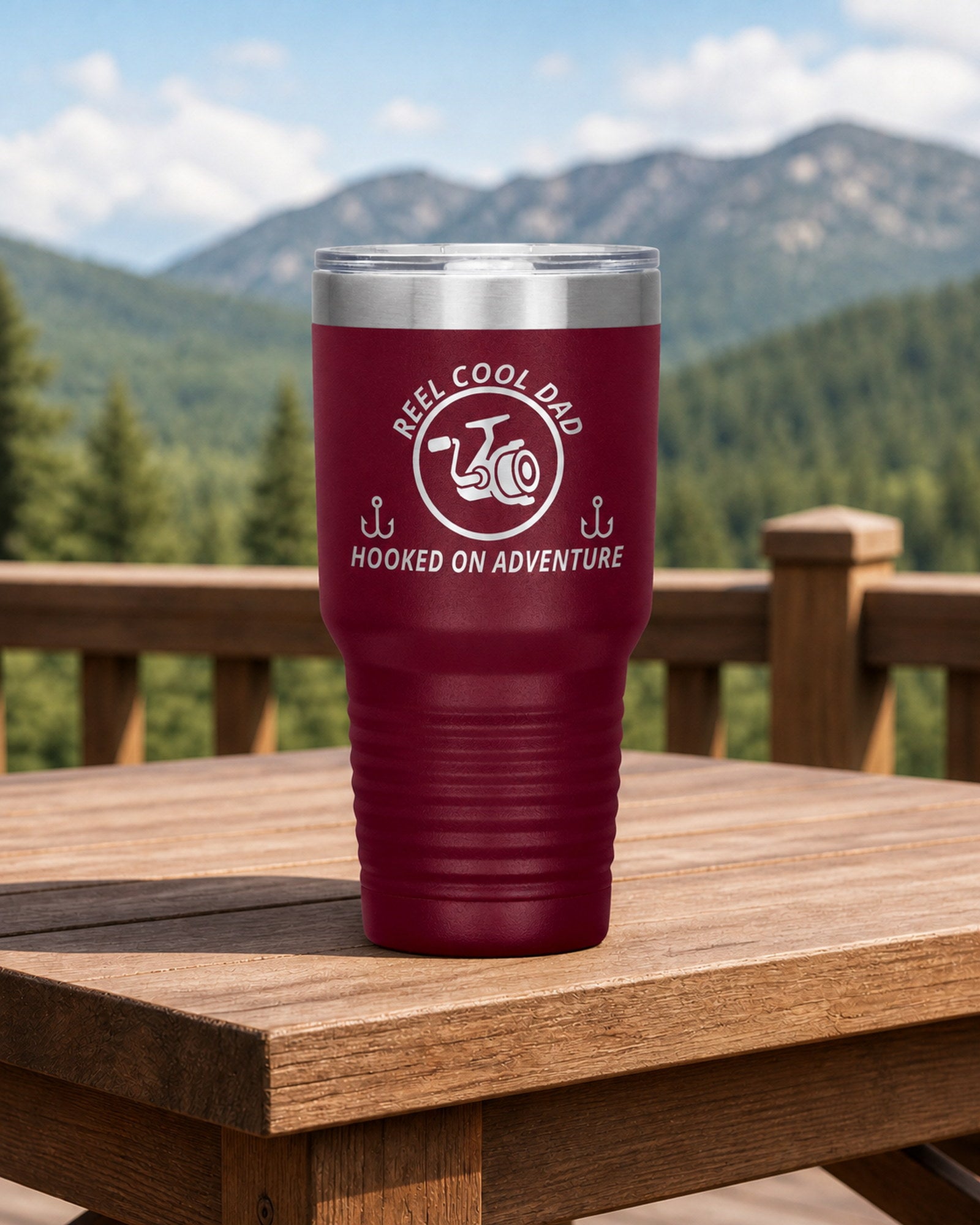 Maroon 30-ounce REEL COOL DAD engraved tumbler on a sturdy cabin porch table overlooking forested mountains.