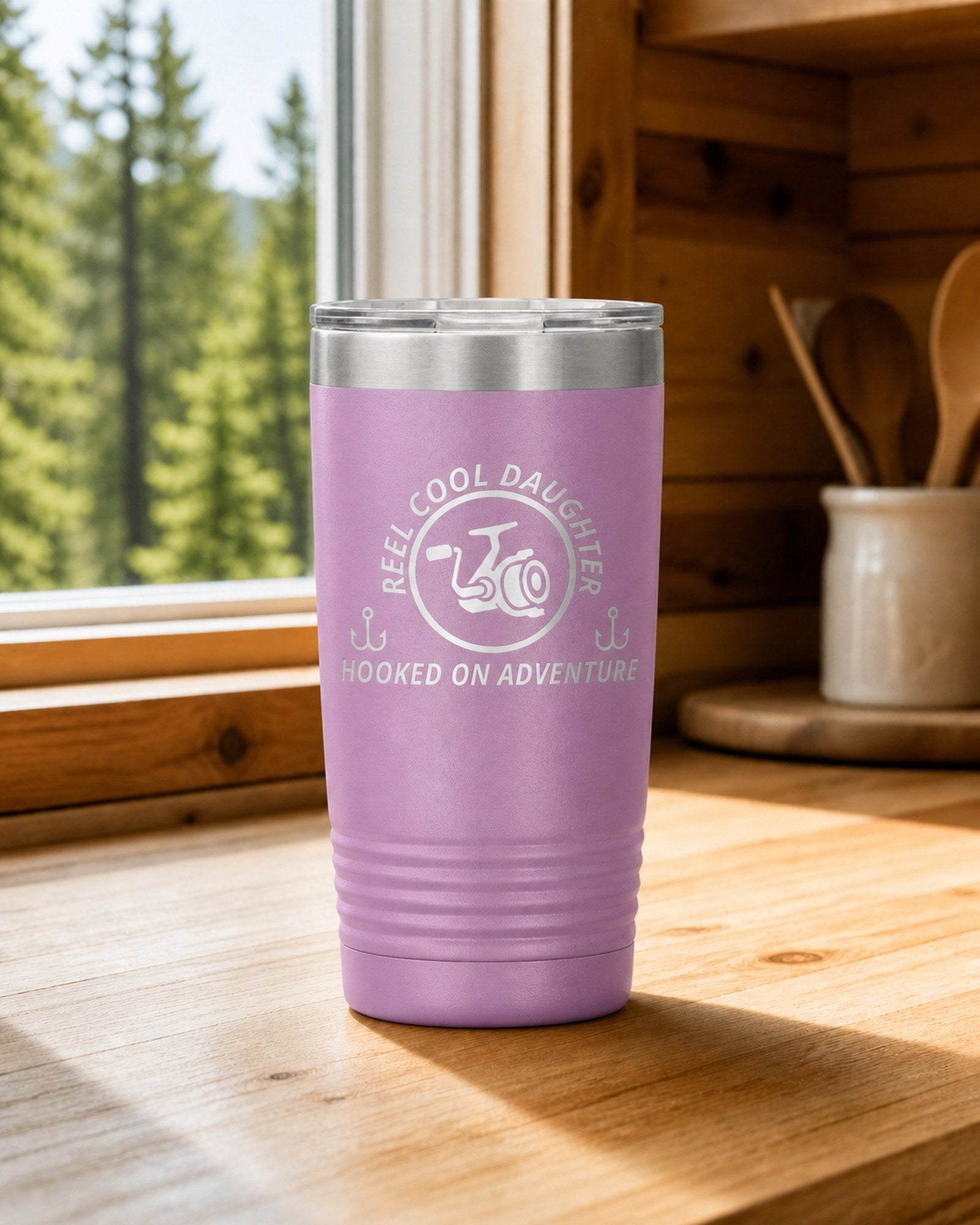 Light Purple 20-ounce REEL COOL DAUGHTER engraved tumbler on a bright wood cabin counter beside a window overlooking green forest.