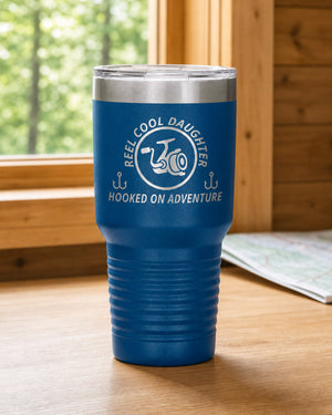 Blue 30-ounce REEL COOL DAUGHTER engraved tumbler on a bright wood cabin counter beside a window overlooking green forest.