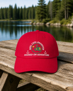 Cranberry REEL COOL DAUGHTER embroidered dad cap on a weathered wooden picnic table beside a pine-lined forest lake.