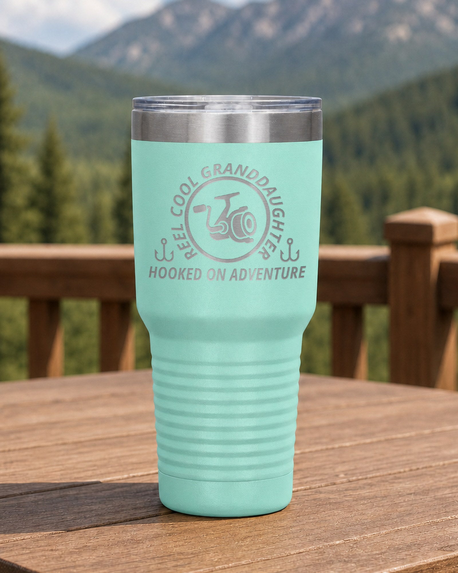 Teal 30-ounce REEL COOL GRANDDAUGHTER engraved tumbler on a sturdy cabin porch table overlooking forested mountains.