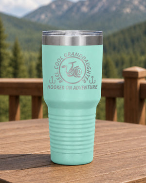 Teal 30-ounce REEL COOL GRANDDAUGHTER engraved tumbler on a sturdy cabin porch table overlooking forested mountains.