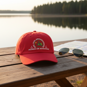 REEL COOL GRANDDAUGHTER cranberry dad cap on a wooden table in a warm lakeside setting.