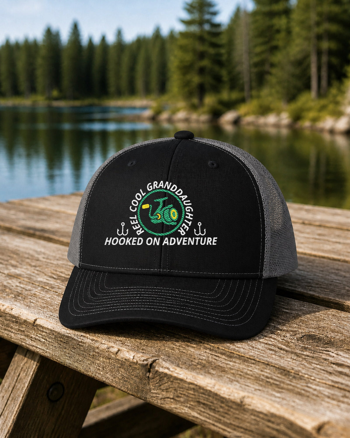 Black and Charcoal REEL COOL GRANDDAUGHTER embroidered trucker cap on a weathered wooden picnic table beside a pine-lined forest lake.