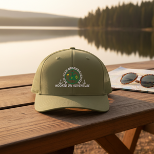 REEL COOL GRANDDAUGHTER trucker cap with loden front and loden mesh back on a wooden table in a warm lakeside setting.