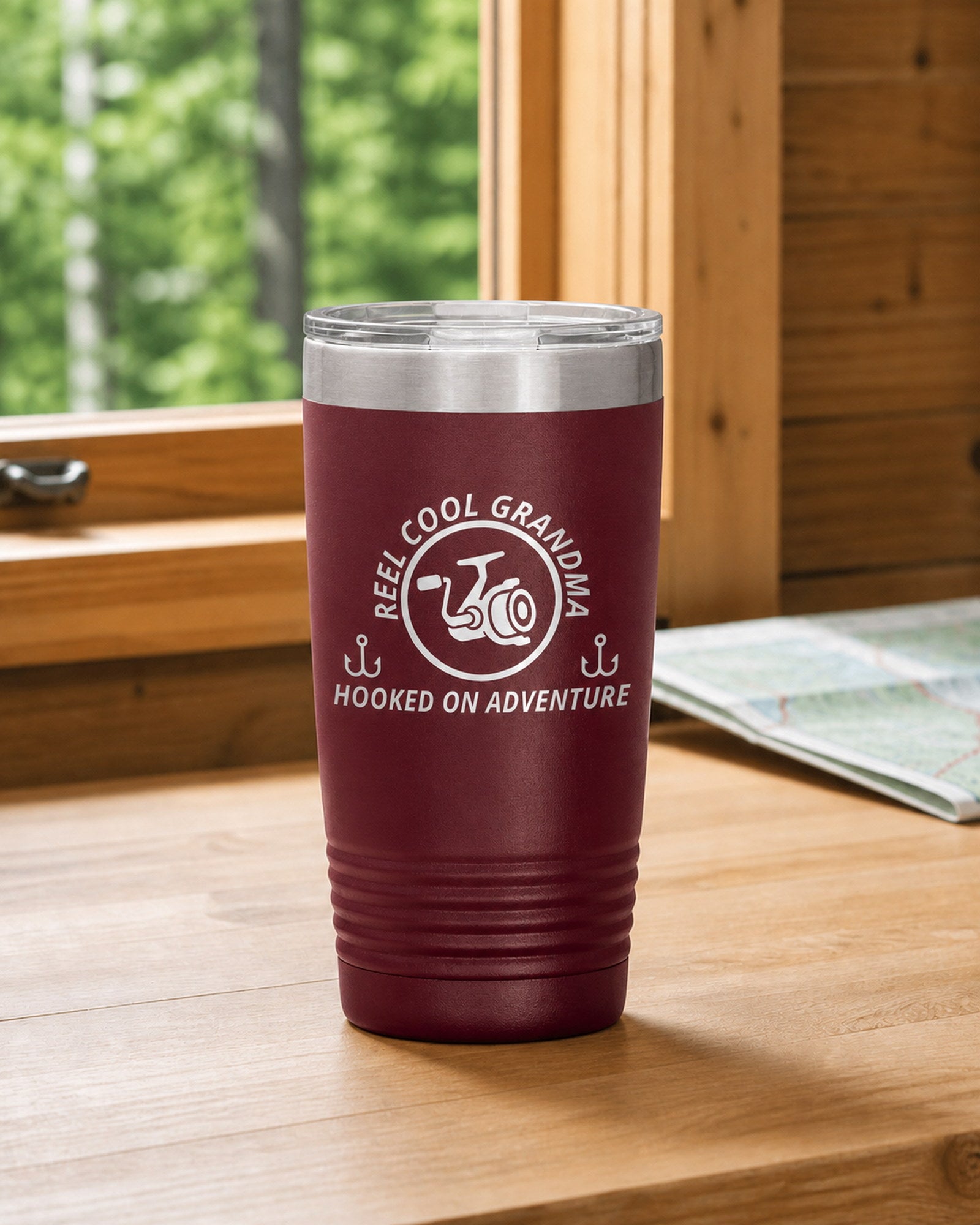 Maroon 20-ounce REEL COOL GRANDMA engraved tumbler on a bright wood cabin counter beside a window overlooking green forest.