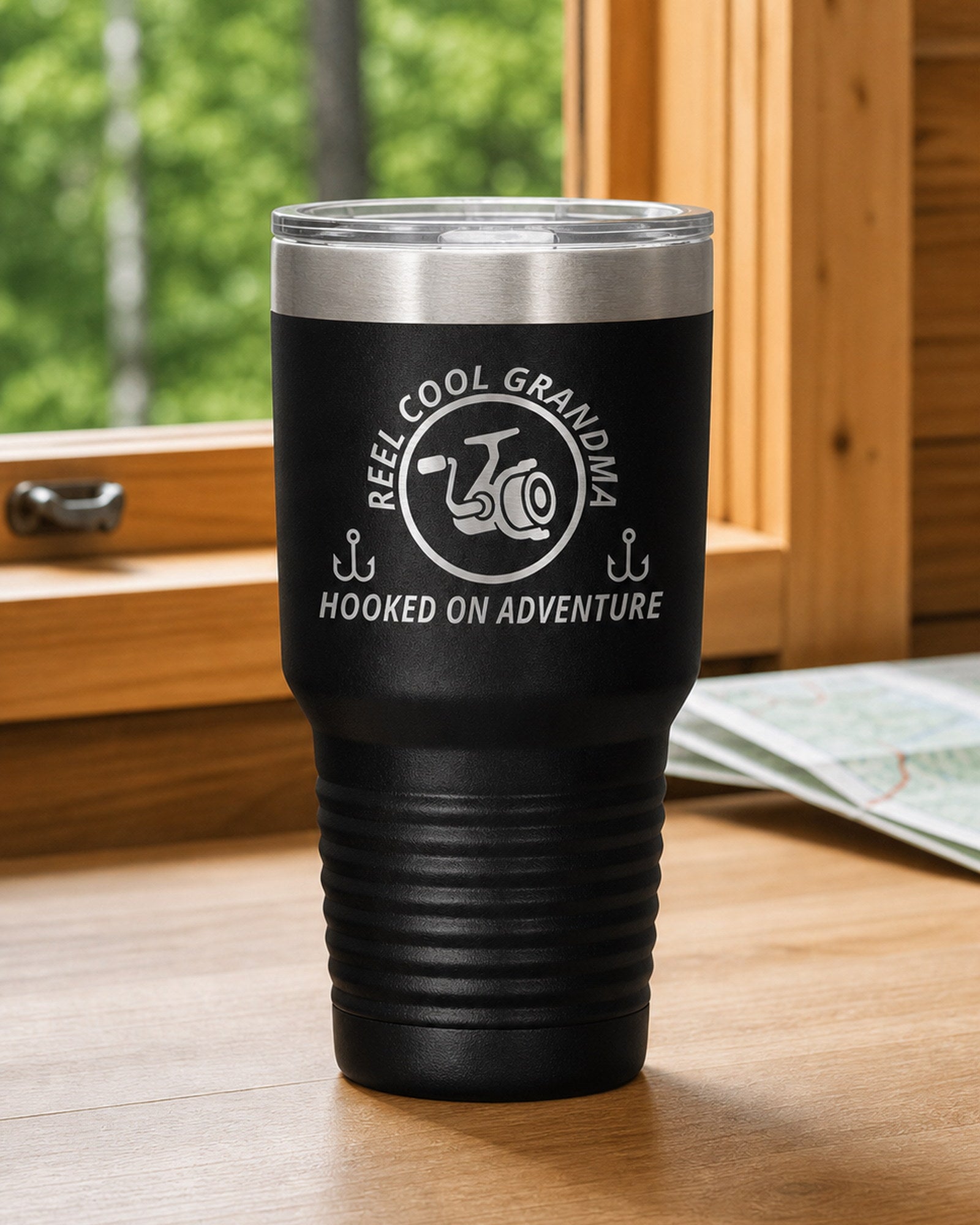 Black 30-ounce REEL COOL GRANDMA engraved tumbler on a bright wood cabin counter beside a window overlooking green forest.