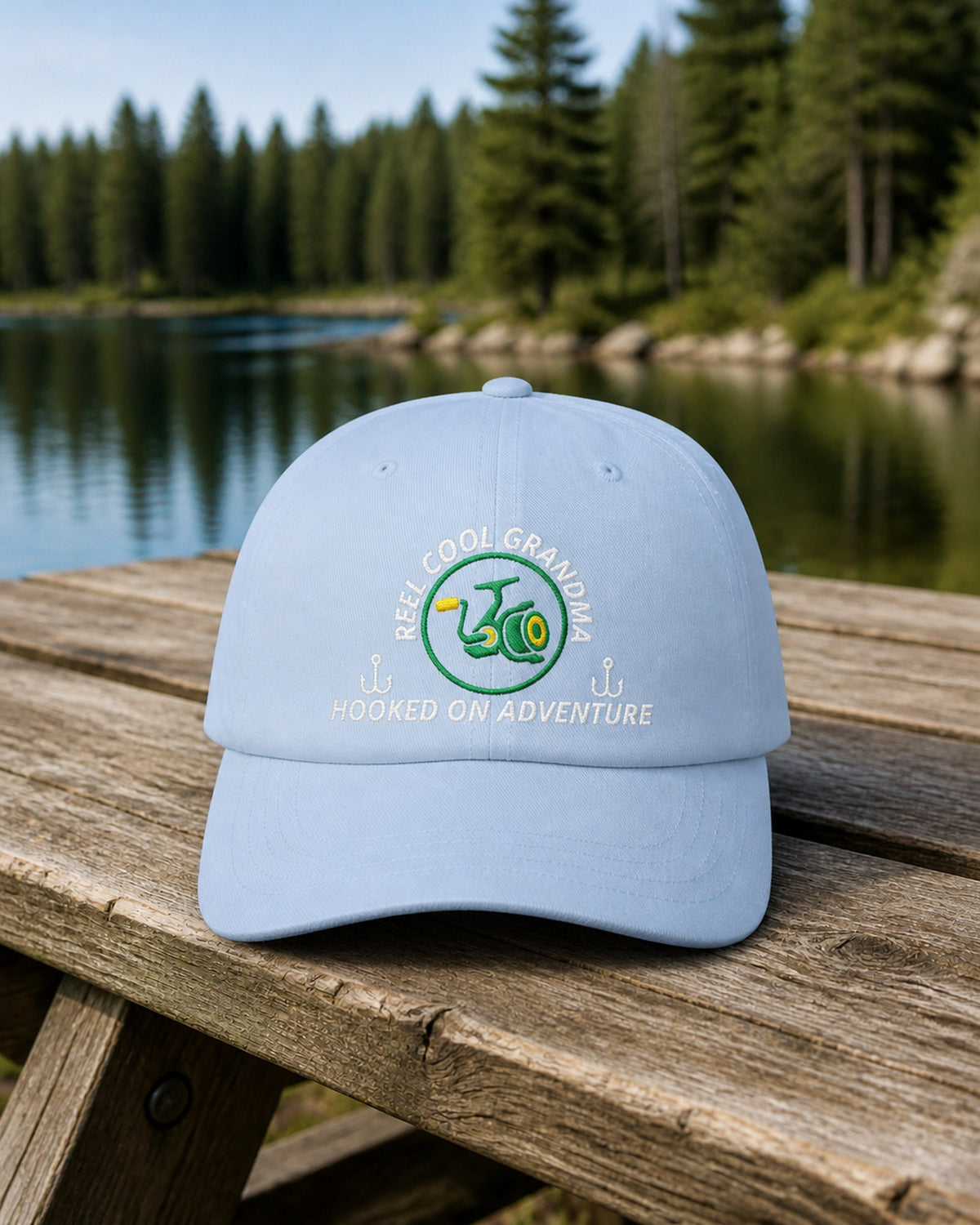 Light Blue REEL COOL GRANDMA embroidered dad cap on a weathered wooden picnic table beside a pine-lined forest lake.