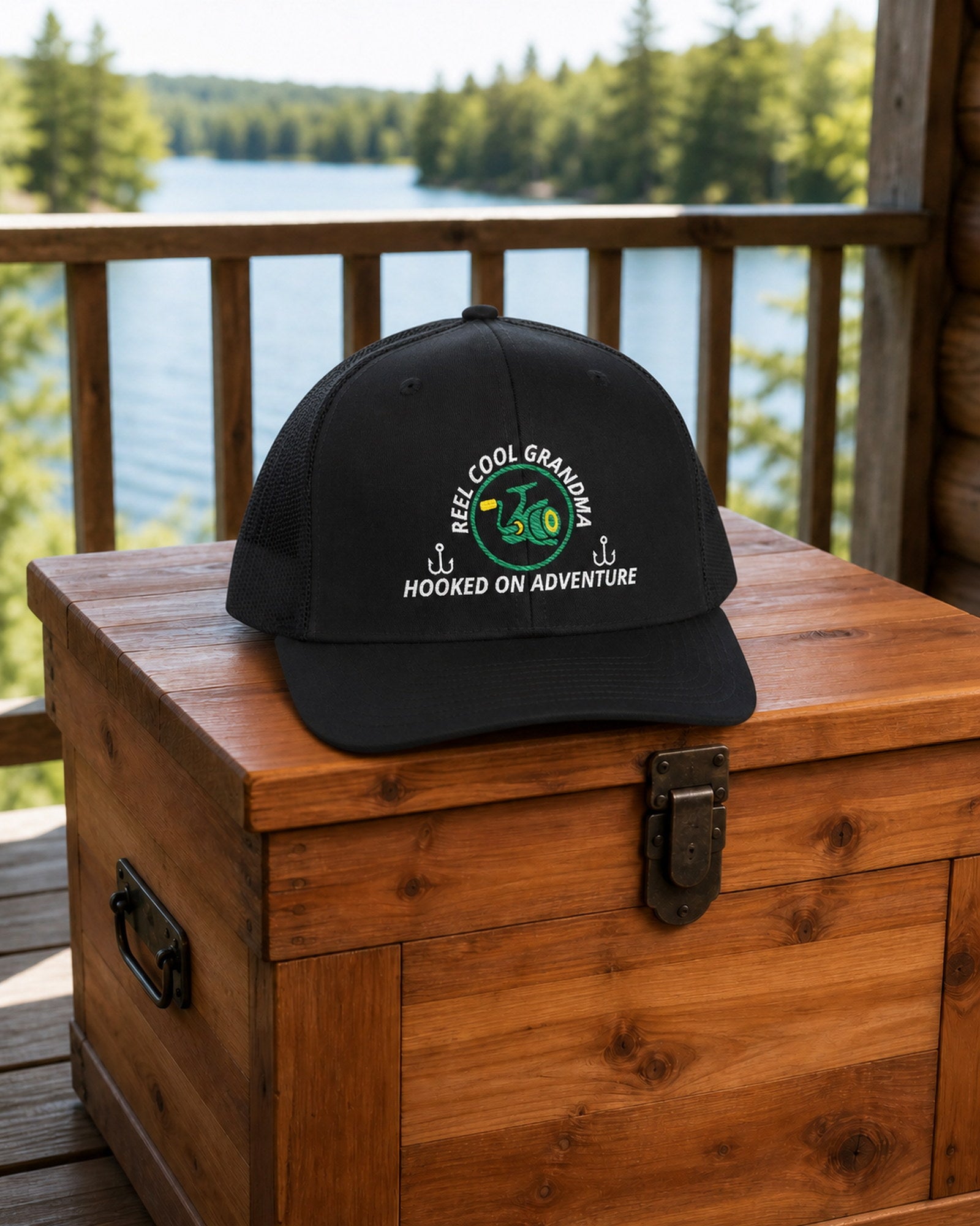 Black REEL COOL GRANDMA embroidered trucker cap on a cedar gear chest overlooking a pine-lined forest lake.