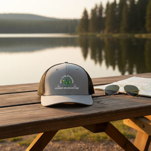 REEL COOL GRANDMA trucker cap with heather gray front and black mesh back on a wooden table in a warm lakeside setting.
