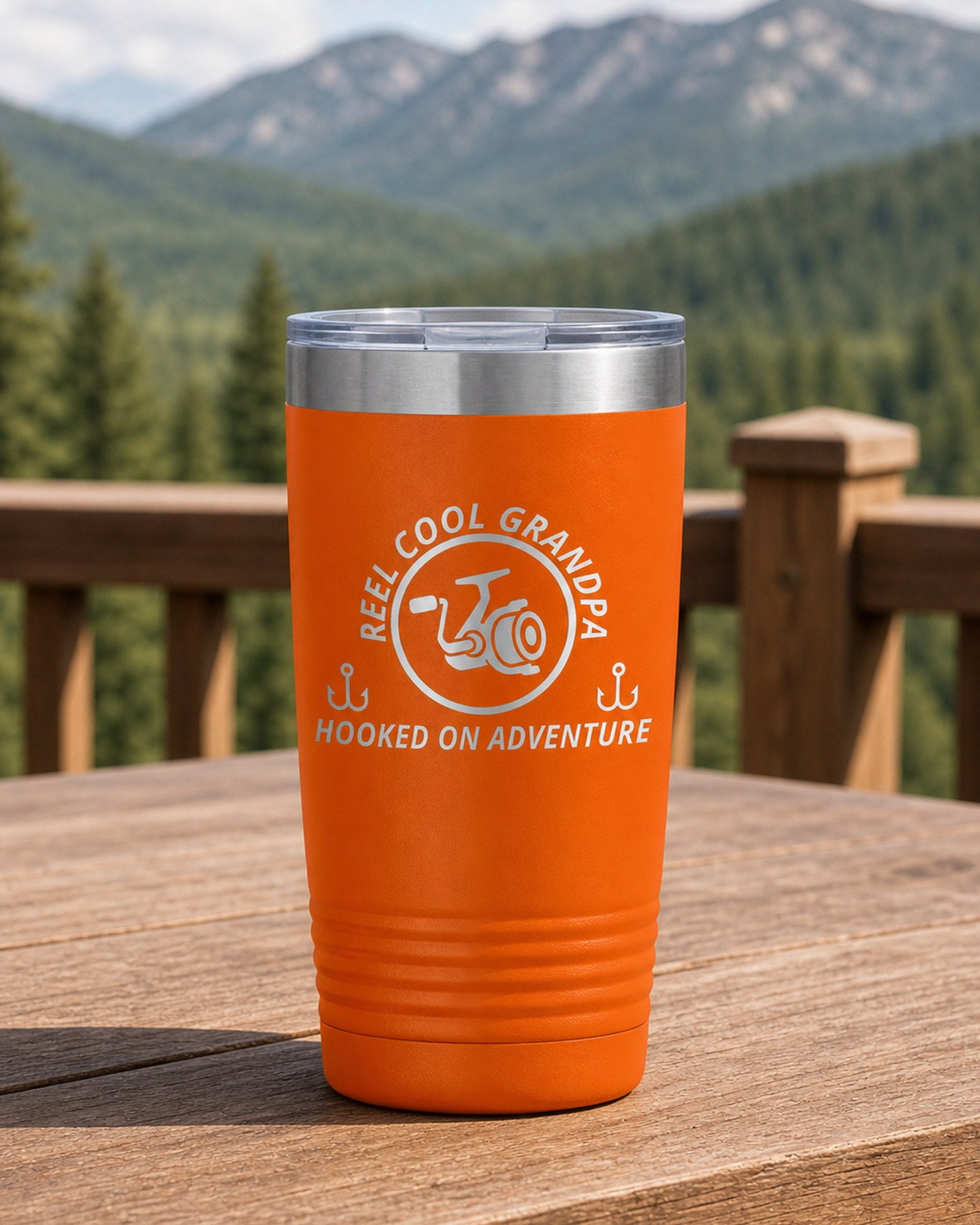 Orange 20-ounce REEL COOL GRANDPA engraved tumbler on a sturdy cabin porch table overlooking forested mountains.