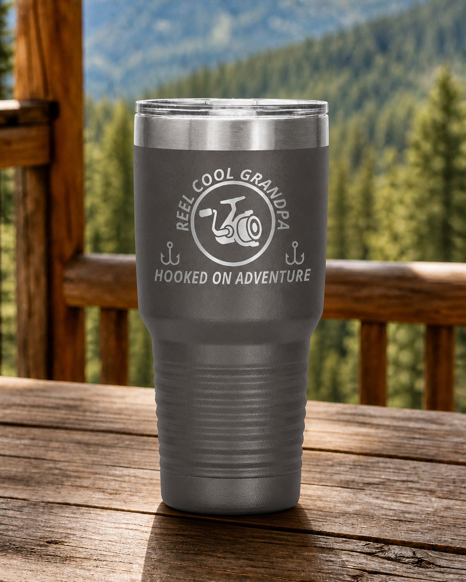 Pewter 30-ounce REEL COOL GRANDPA engraved tumbler on a sturdy cabin porch table overlooking forested mountains.