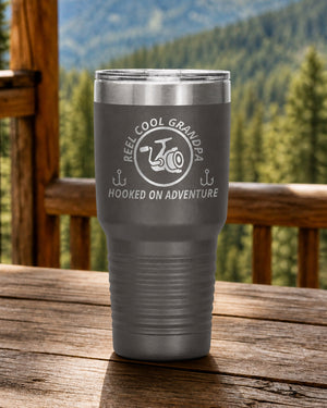 Pewter 30-ounce REEL COOL GRANDPA engraved tumbler on a sturdy cabin porch table overlooking forested mountains.