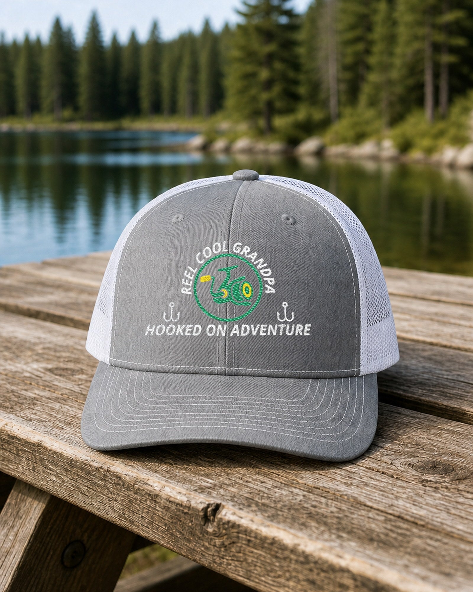Heather Grey and White REEL COOL GRANDPA embroidered trucker cap on a weathered wooden picnic table beside a pine-lined forest lake.