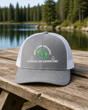 Heather Grey and White REEL COOL GRANDPA embroidered trucker cap on a weathered wooden picnic table beside a pine-lined forest lake.