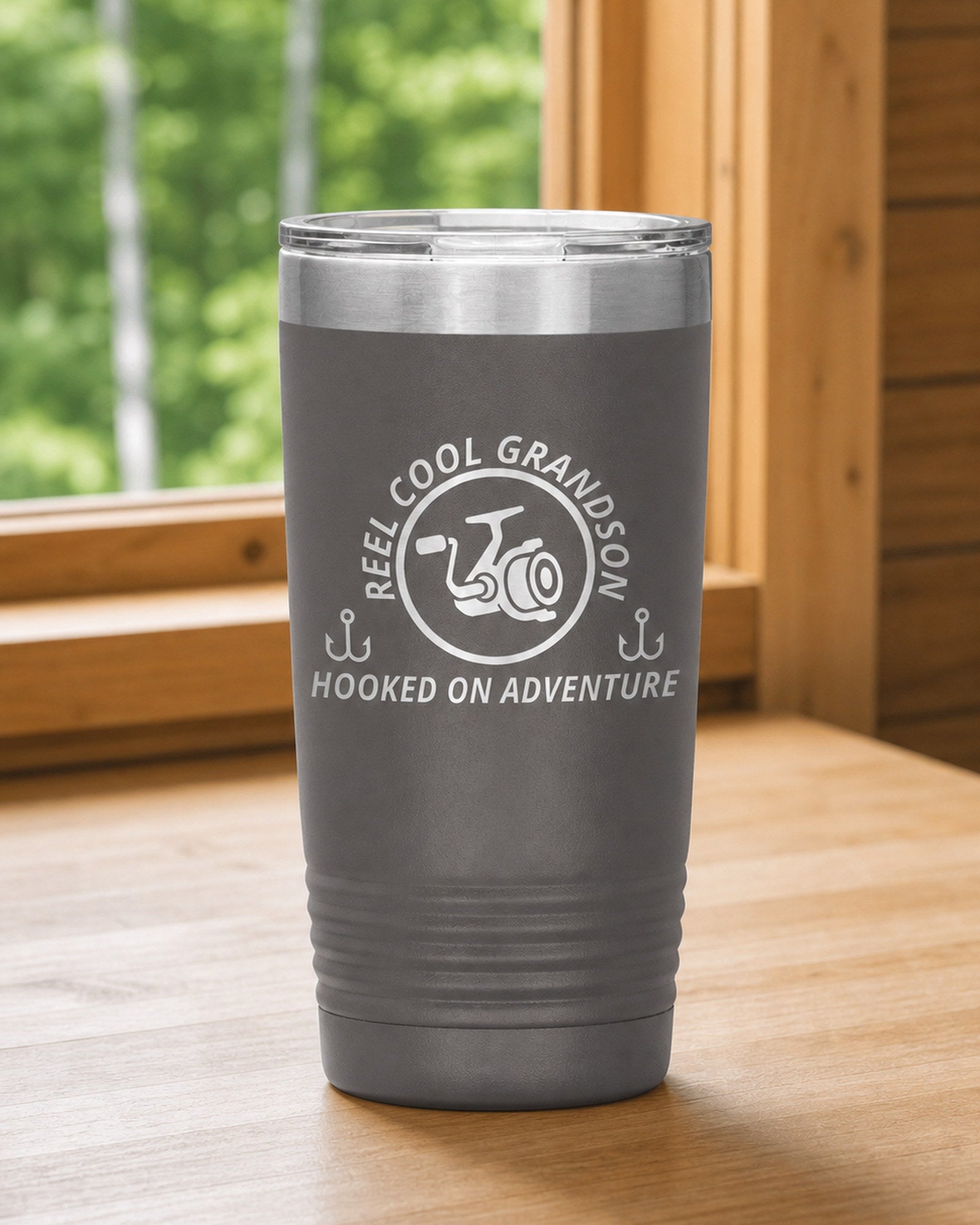 Pewter 20-ounce REEL COOL GRANDSON engraved tumbler on a bright wood cabin counter beside a window overlooking green forest.