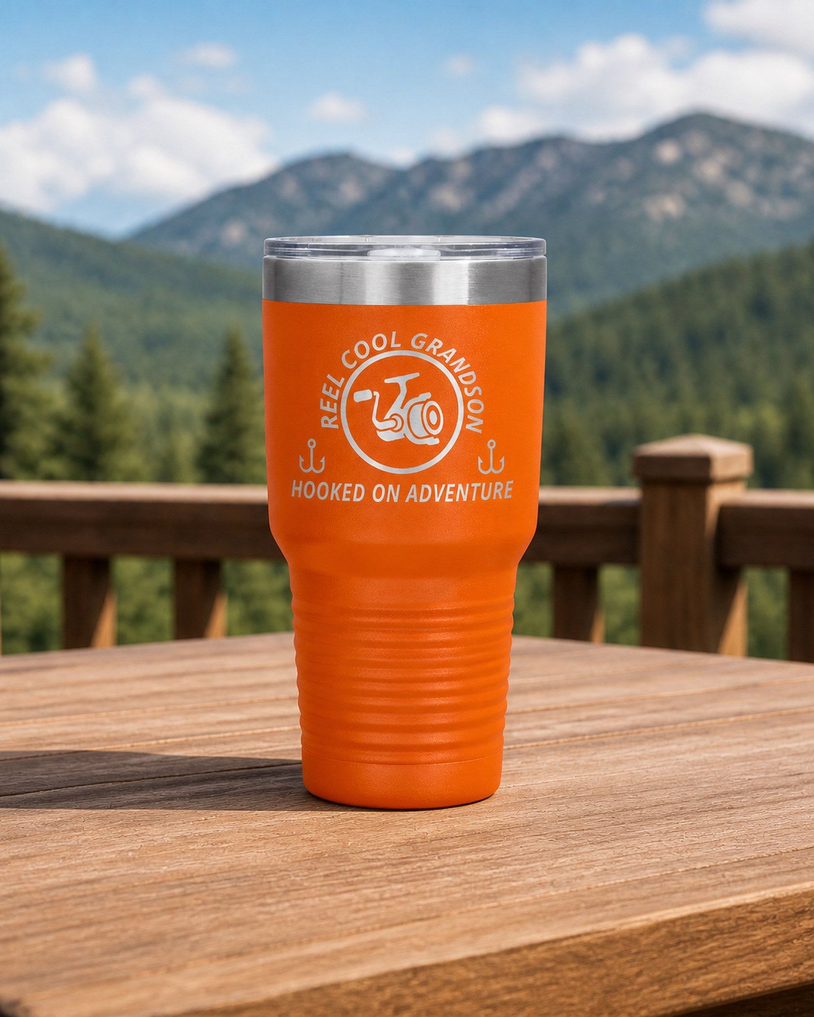 Orange 30-ounce REEL COOL GRANDSON engraved tumbler on a sturdy cabin porch table overlooking forested mountains.