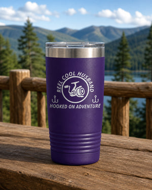 Purple 20-ounce REEL COOL HUSBAND engraved tumbler on a sturdy cabin porch table overlooking forested mountains.