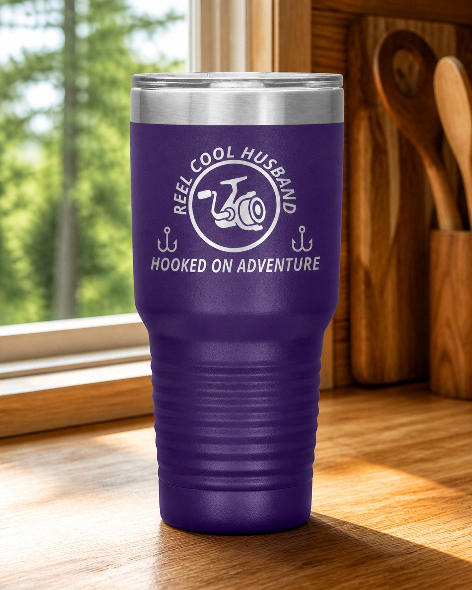 Purple 30-ounce REEL COOL HUSBAND engraved tumbler on a bright wood cabin counter beside a window overlooking green forest.