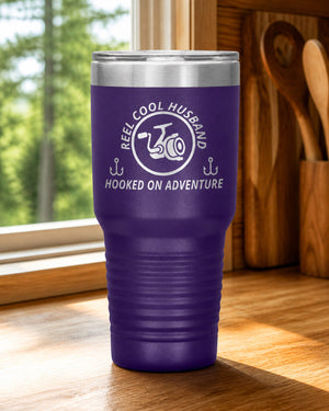 Purple 30-ounce REEL COOL HUSBAND engraved tumbler on a bright wood cabin counter beside a window overlooking green forest.