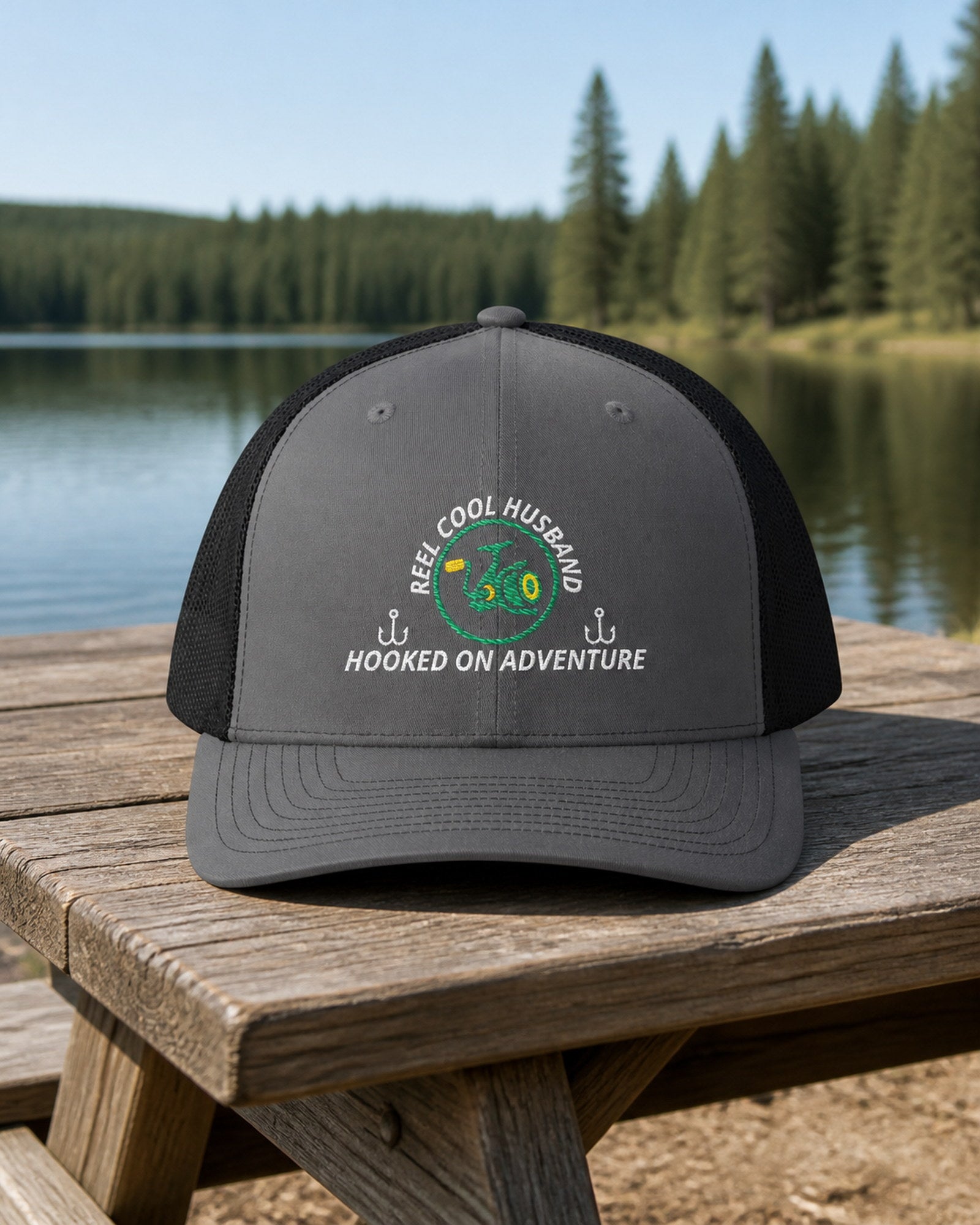 Charcoal and Black REEL COOL HUSBAND embroidered trucker cap on a weathered wooden picnic table beside a pine-lined forest lake.