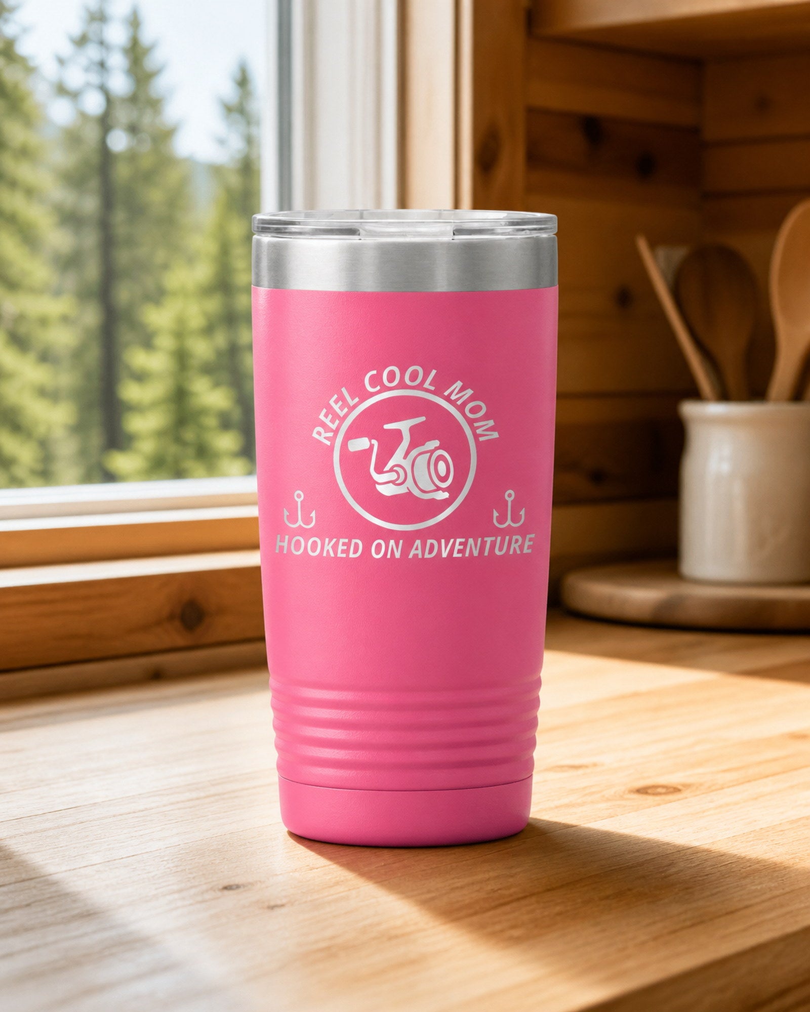 Pink 20-ounce REEL COOL MOM engraved tumbler on a bright wood cabin counter beside a window overlooking green forest.