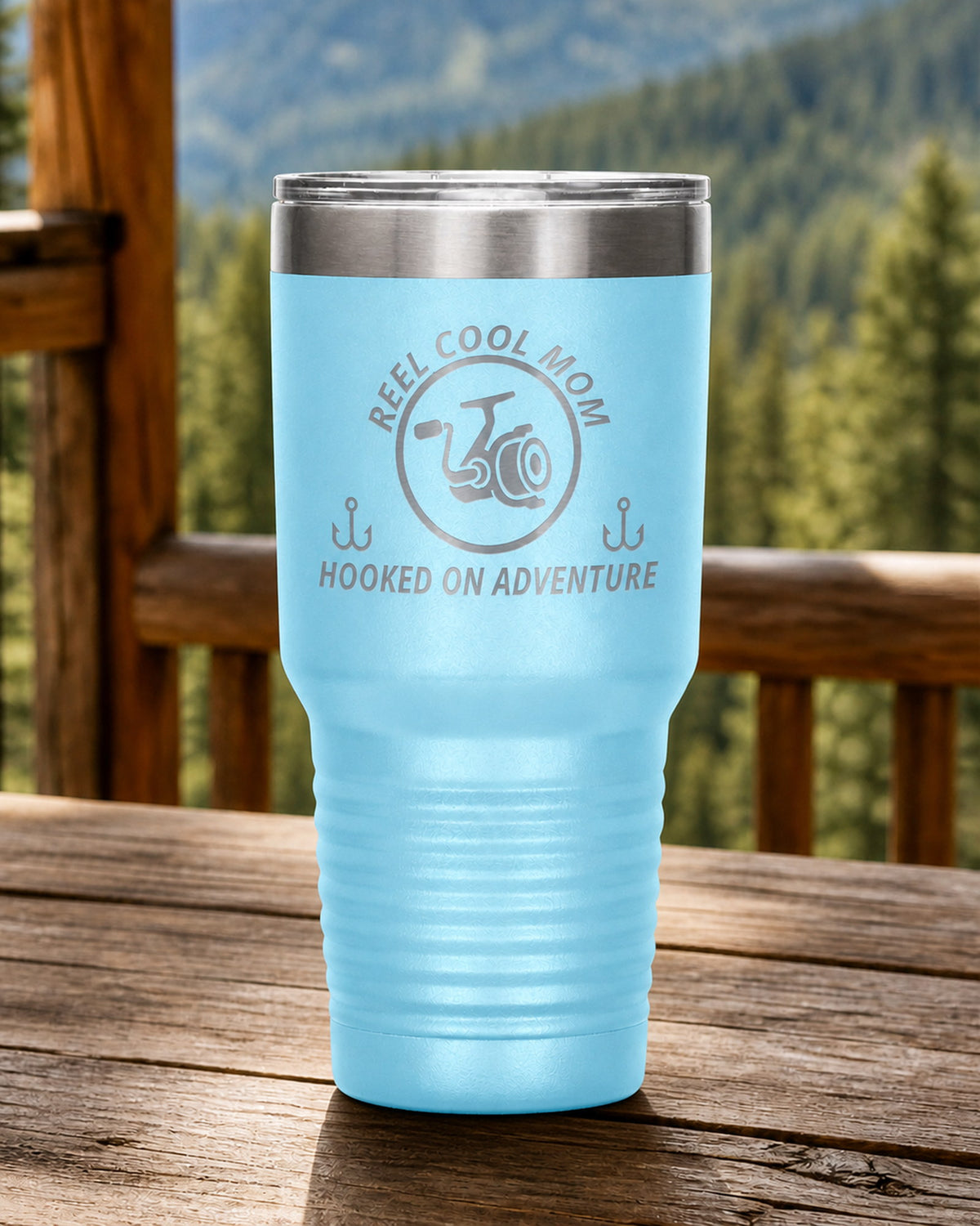 Light Blue 30-ounce REEL COOL MOM engraved tumbler on a sturdy cabin porch table overlooking forested mountains.