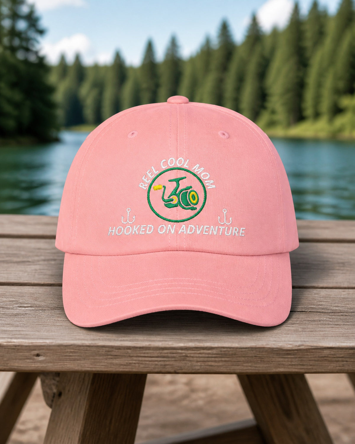 Pink REEL COOL MOM embroidered dad cap on a weathered wooden picnic table beside a pine-lined forest lake.