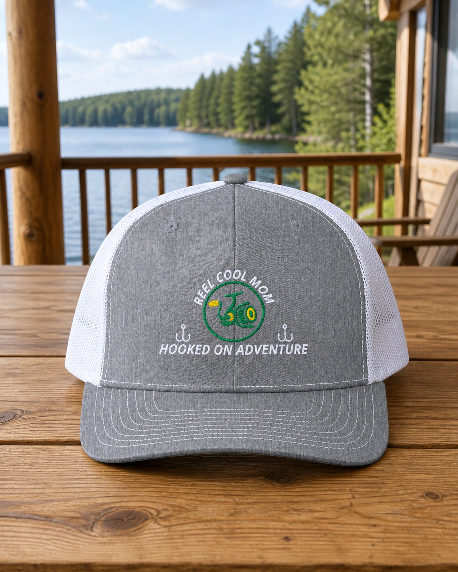 Heather Grey and White REEL COOL MOM embroidered trucker cap on a full-size cedar cabin porch table overlooking a forest lake.