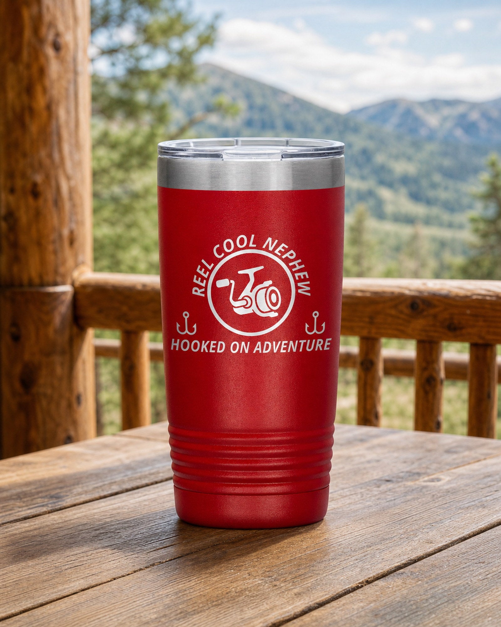 Red 20-ounce REEL COOL NEPHEW engraved tumbler on a sturdy cabin porch table overlooking forested mountains.