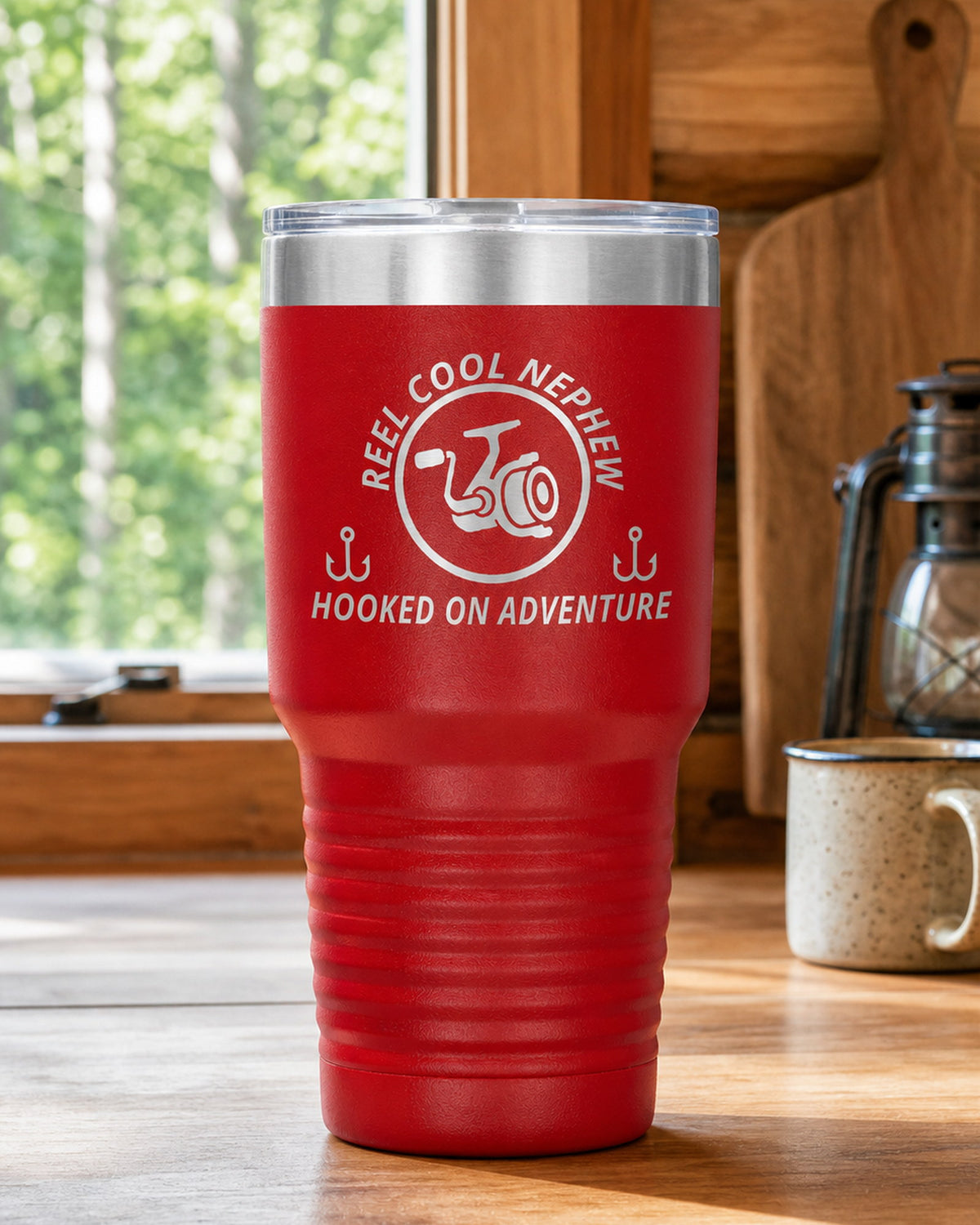 Red 30-ounce REEL COOL NEPHEW engraved tumbler on a bright wood cabin counter beside a window overlooking green forest.