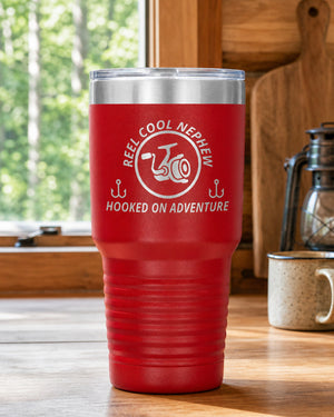 Red 30-ounce REEL COOL NEPHEW engraved tumbler on a bright wood cabin counter beside a window overlooking green forest.