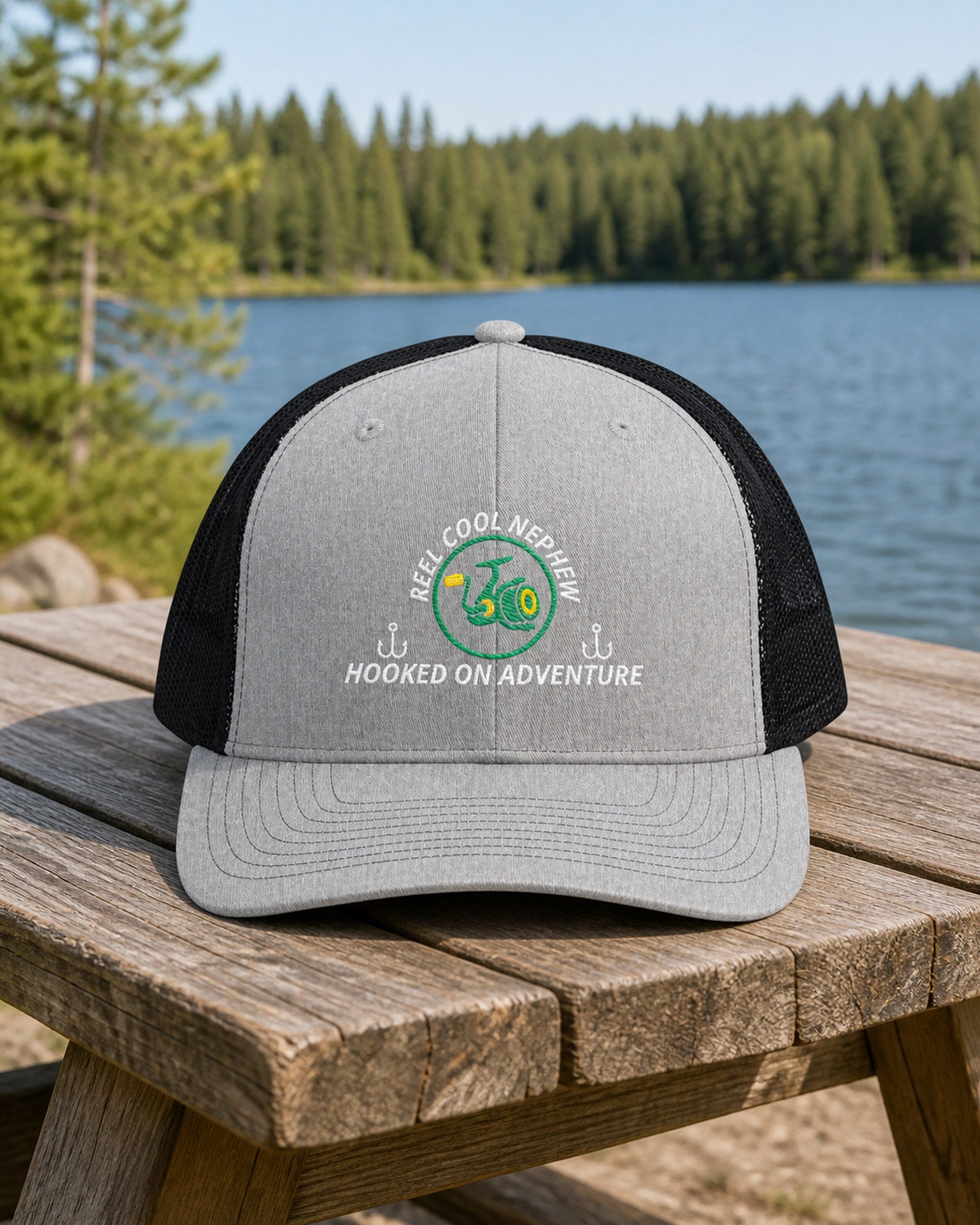 Heather Grey and Black REEL COOL NEPHEW embroidered trucker cap on a weathered wooden picnic table beside a pine-lined forest lake.