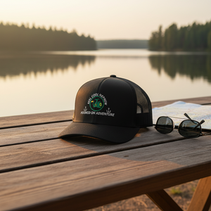 REEL COOL NEPHEW trucker cap with black front and black mesh back on a wooden table in a warm lakeside setting.
